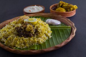 Kolkata style mutton biryani with potato and egg served on clay plate and banana leaf with mutton curry and curd raita. shot against black background.